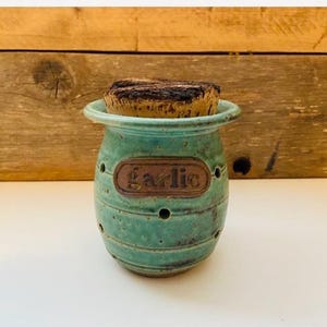 May include: A turquoise ceramic garlic keeper with a wide rim and a cork lid. The vessel features horizontal ridges, small ventilation holes around the body, and a central rectangular plaque with the word garlic embossed in dark brown lettering.