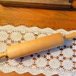 May include: A light-colored wooden rolling pin with rounded handles, resting on a white lace doily with a floral pattern. The rolling pin is a kitchen utensil for baking.