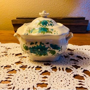 May include: A white ceramic lidded serving dish with teal floral designs. The dish has two handles and sits on a white lace doily. The background includes a wooden box and a light-colored wall.