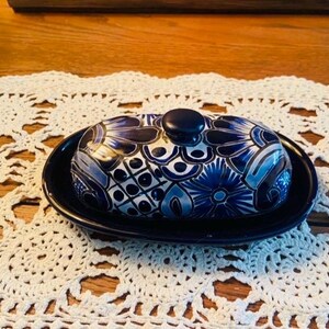 May include: A ceramic butter dish with a lid, featuring a dark blue base and lid. The lid is adorned with a detailed, hand-painted design in shades of blue and white, with a small knob on top. The dish sits on a dark blue oval base.