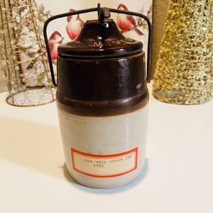 May include: A vintage stoneware crock jar with a dark brown lid and handle. The jar has a light beige base with a dark brown band. A rectangular label reads "the weir crock jar 1892". The jar is a decorative kitchen item.