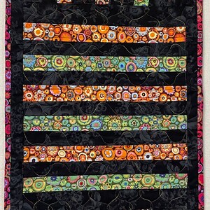 May include: A black quilted table runner with a colorful geometric pattern. The pattern features circles and stripes in shades of orange, red, green, and blue. The runner is edged with a black border.