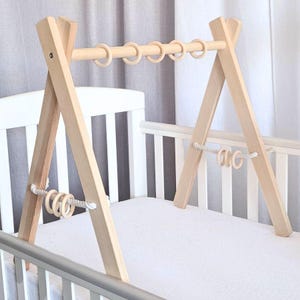 Pode incluir: Um ginásio de brincar para bebés de madeira com acabamento natural. O ginásio tem uma barra horizontal com anéis de madeira e uma corda com anéis de madeira pendurados nela. O ginásio está instalado sobre um berço branco.
