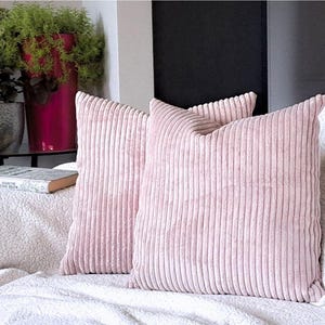 May include: Three pink corduroy throw pillows with a ribbed texture. The pillows are arranged on a white couch with a white throw blanket.