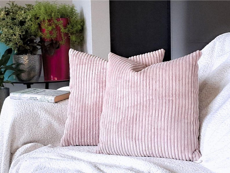 May include: Three pink corduroy throw pillows with a ribbed texture. The pillows are arranged on a white couch with a white throw blanket.