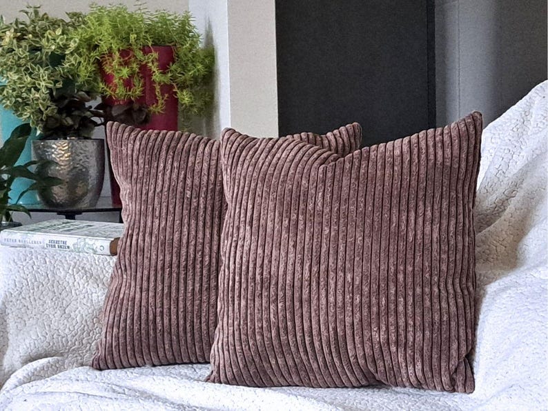 May include: Two brown corduroy throw pillows with a vertical ribbed texture. The pillows are on a white couch.
