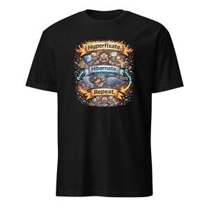 May include: Black t-shirt with a vibrant graphic. The design includes the words "Hyperfixate," "Hibernate," and "Repeat," accompanied by illustrations of people using technology and sleeping. The design is framed by flames and small icons.
