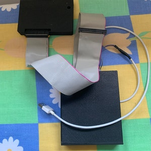 Puede incluir: Un componente de computadora negro con un cable de cinta conectado a él. El cable está conectado a un segundo componente negro. Un cable USB blanco también está conectado al segundo componente.