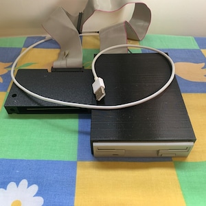 Puede incluir: Una unidad de disquete en blanco y negro con un cable USB conectado. La unidad está sentada sobre una tela a cuadros de colores.