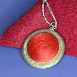 Op de afbeelding: Een ronde, rode hanger met een textuuroppervlak, gezet in een goudkleurige lijst. Hij hangt aan een zilverkleurig, gevlochten koord. De hanger rust op een rode viltachtergrond op een blauw oppervlak.