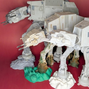 Peut inclure: Une collection de quatre figurines articulées de marcheurs en plastique gris et blanc avec des canons montés sur la tête. Chaque figurine repose sur une base unique, incluant de la roche grise texturée, un terrain vert vif et de la neige blanche.