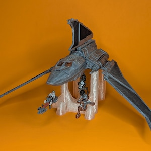 Könnte beinhalten: Ein graues und schwarzes Raumschiffmodell mit einem langen, dünnen Flügel und einem großen, flachen Flügel. Das Raumschiff steht auf einem weißen Ständer mit drei Figuren darauf.