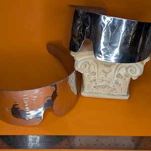 May include: Two pairs of futuristic silver sunglasses with a hammered metal texture. One pair rests on a decorative white column, while the other lies flat. A stainless steel ruler is at the bottom of the image.