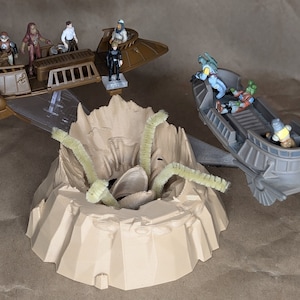 May include: A Star Wars toy playset featuring miniature vehicles and figures. The scene includes a brown vehicle with several figures, a gray vehicle with figures, and a beige terrain piece with green pipe cleaner-like elements.