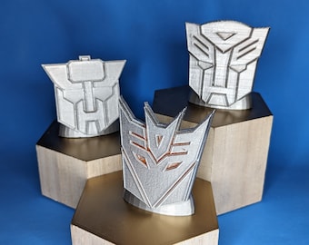 May include: Three silver-colored Transformers symbols, each mounted on a hexagonal wooden base. The symbols are the Autobot and Decepticon insignias, and a third, similar design. The background is a solid blue.