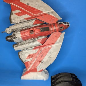 Puede incluir: Un modelo de nave espacial rojo y blanco con un soporte negro. La nave espacial tiene un diseño curvo y está detallada con rayas rojas y blancas. El soporte es de plástico y tiene una superficie texturizada.