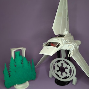 May include: A white model of a Star Wars Snowspeeder with a red cockpit window. The Snowspeeder is on a stand with a white Imperial symbol. A green model of a forest is in the background.
