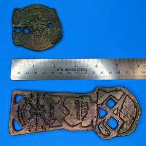 Two metal objects with a dark brown and green color. The objects are on a blue surface with a ruler showing their comparative sizes. The larger object is shaped like a skull with a hole in the middle. The smaller object is round with a hole in the center and a cross design.
