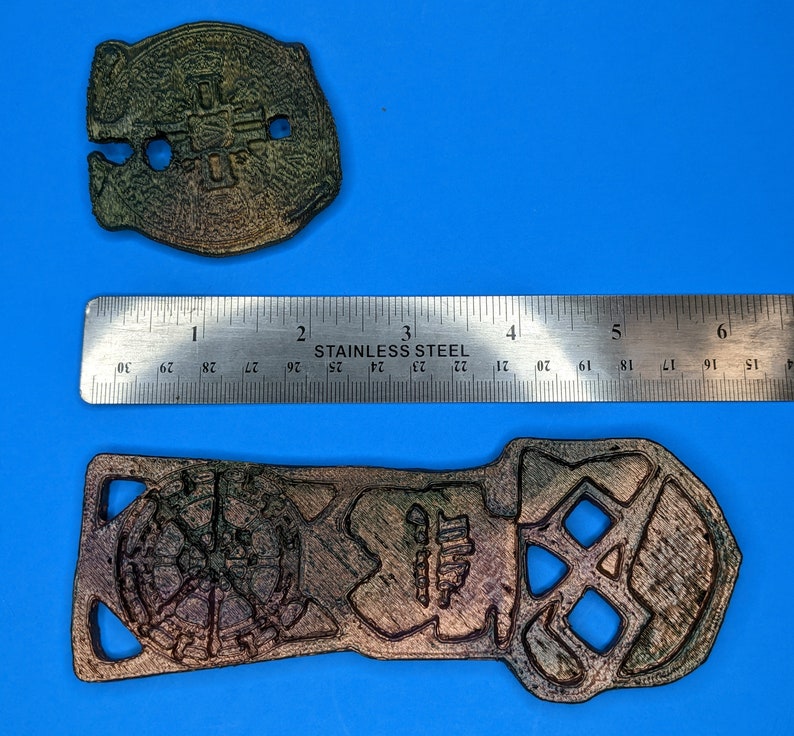 Two metal objects with a dark brown and green color. The objects are on a blue surface with a ruler showing their comparative sizes. The larger object is shaped like a skull with a hole in the middle. The smaller object is round with a hole in the center and a cross design.