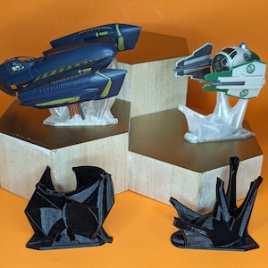 May include: Three 3D printed spaceship models on display stands. The spaceships are painted in blue, green, and white. The stands are made of clear plastic. The spaceships are on a wooden platform.