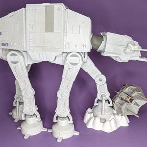 Puede incluir: Un juguete grande de caminante AT-AT blanco con cuerpo rectangular, cuatro patas articuladas con juntas circulares y una cabeza con cañones dobles. Un juguete de snowspeeder blanco más pequeño está colocado en el suelo junto a uno de los pies.