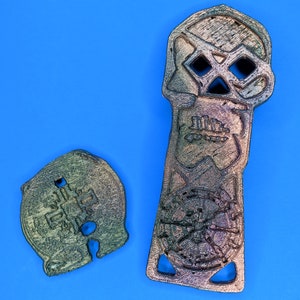 Two 3D printed objects, one is a skull shaped object with a circular design in the center and the other is a round object with a cross design in the center. Both objects are metallic with distressed weathering paint.
