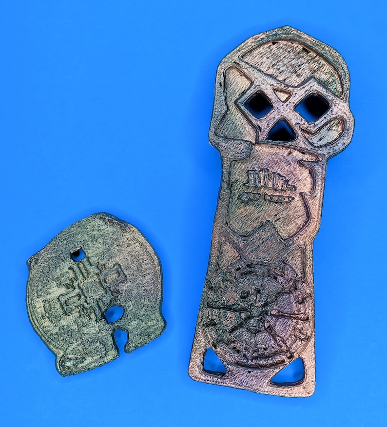 Two 3D printed objects, one is a skull shaped object with a circular design in the center and the other is a round object with a cross design in the center. Both objects are metallic with distressed weathering paint.
