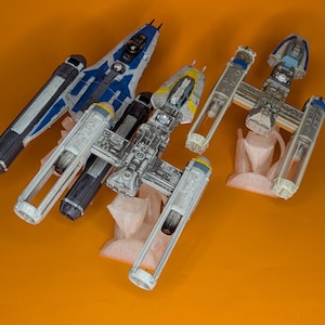 May include: Three miniature starfighter models displayed on translucent stands. One model is blue and white, while the other two are grey and yellow with exposed engine structures. The models feature detailed cockpits and mechanical components.