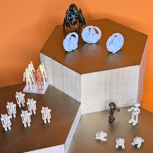 May include: A collection of miniature figurines, including robots, skeletons, and other sci-fi themed characters. The figurines are in various colors, including black, white, and brown. The figurines are arranged on a multi-level display with an orange background.