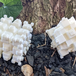 May include: Two white 3D printed geometric shapes, resembling cubes, are placed on a bed of black mulch. The shapes are made up of many smaller cubes, creating a complex and intricate design.