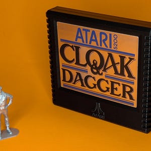 May include: A black Atari 5200 video game cartridge for the game Cloak & Dagger with a gold label and black text, positioned next to a small, silver-colored metal figurine of a person holding a handgun, both set against a solid orange background.