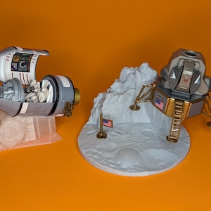 May include: A detailed model of a spacecraft and a lunar module, in shades of grey, white, and gold. The spacecraft has an open hatch revealing astronaut figures. The lunar module features a ladder and an American flag. The models are set on a grey lunar surface.