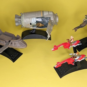 May include: Five Star Wars toy vehicles on black display stands. The vehicles include a brown speeder, a red speeder, a gray and orange spaceship, and two brown and green speeder bikes.