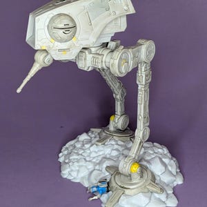 May include: A detailed model of an AT-ST walker from Star Wars, painted in shades of gray with yellow accents. The model stands on a textured, white base, with a small blue vehicle nearby. The walker has articulated legs and a detailed head.
