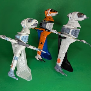 May include: Three toy spacecraft models in white, orange, and blue, with detailed wings and cockpit designs. The models are displayed on stands against a green background, showcasing their intricate features and color schemes. The toys are likely from a science fiction theme.