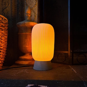 Puede incluir: Lámpara de mesa cilíndrica con pantalla acanalada verticalmente que emite una luz amarilla cálida. La lámpara se apoya sobre una base de hormigón gris. Diseño minimalista, adecuado para la decoración del hogar. Altura aproximada 25 cm.