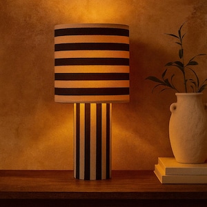 Puede incluir: Una lámpara de mesa con base y pantalla cilíndricas. La base tiene rayas verticales negras y crema, mientras que la pantalla presenta rayas horizontales negras. La lámpara está sobre una superficie de madera, con un jarrón y libros al fondo.