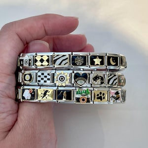 Puede incluir: Tres pulseras de abalorios plateadas con varios abalorios negros y dorados. Los abalorios incluyen una cámara, una estrella, una huella de pata, un gato y un corazón con un ojo. Las pulseras se sostienen en una mano.