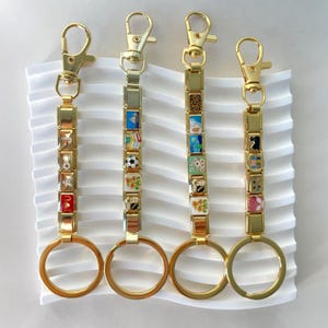 Peut inclure: Quatre porte-clés dorés avec des breloques décoratives. Chaque porte-clés comprend un fermoir, une série de breloques carrées avec divers motifs et un grand anneau. Les breloques présentent des lettres, des chiffres et de petites illustrations multicolores.