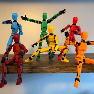 May include: Six articulated action figures in various poses, each a different color: blue, green, red, yellow, and orange. The figures are posed on a wooden shelf, with arms and legs in different positions.