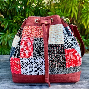 May include: A patchwork bucket bag with a drawstring closure and a shoulder strap. The bag features a variety of fabric patterns in red, black, white, and cream. The bag's trim and strap are a solid burgundy color. The bag has gold-colored metal hardware.