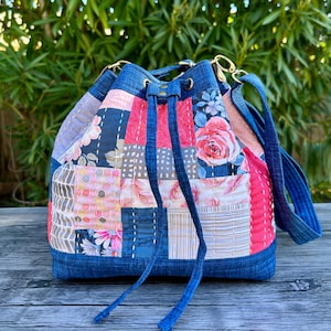 May include: A patchwork bucket bag with a denim base and drawstring closure. The bag features a variety of fabric patterns, including floral and geometric designs in pink, blue, and neutral tones. It has a blue shoulder strap and gold-colored hardware.