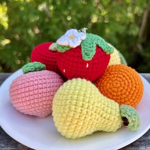 May include: A collection of colorful, crocheted fruit on a white plate. The assortment includes a red strawberry with a white flower, a pink apple, an orange, a yellow pear, and a red fruit. The fruit is arranged on a white plate.