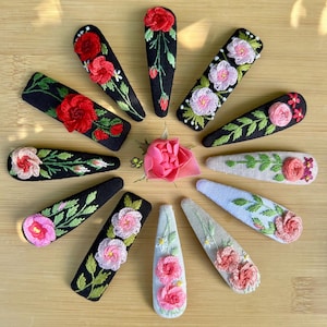 May include: A collection of embroidered hair clips featuring floral designs. The clips showcase various flowers in shades of pink and red, with green leaves, on black, white, and light blue backgrounds. The clips are arranged on a wooden surface.