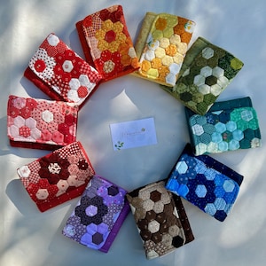 May include: A collection of nine rectangular fabric wallets arranged in a circle, each featuring a quilted hexagonal patchwork design in various colors including red, orange, yellow, green, teal, blue, purple, and brown, with a central card.