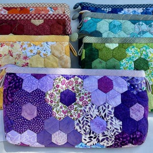 May include: A collection of handmade fabric pouches in various colors and patterns. The pouches feature a hexagonal patchwork design, with colors including yellow, purple, green, and red. Each pouch has a zipper closure and a small handle.