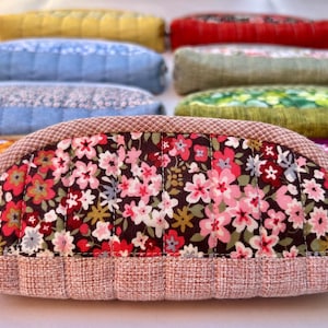 K&ouml;nnte beinhalten: Eine Sammlung halbkreisf&ouml;rmiger, gesteppter Taschen, die in Reihen angeordnet sind. Die vordere Tasche zeigt einen dunkelbraunen Stoff mit einem rosa, roten und wei&szlig;en Blumenmuster, eingefasst von einem strukturierten hellrosa Stoff. Weitere Taschen zeigen verschiedene Farben.