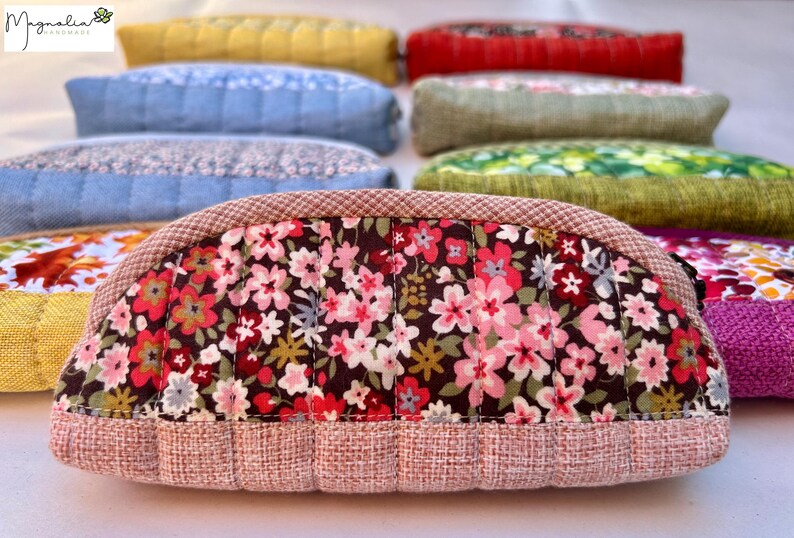 K&ouml;nnte beinhalten: Eine Sammlung halbkreisf&ouml;rmiger, gesteppter Taschen, die in Reihen angeordnet sind. Die vordere Tasche zeigt einen dunkelbraunen Stoff mit einem rosa, roten und wei&szlig;en Blumenmuster, eingefasst von einem strukturierten hellrosa Stoff. Weitere Taschen zeigen verschiedene Farben.