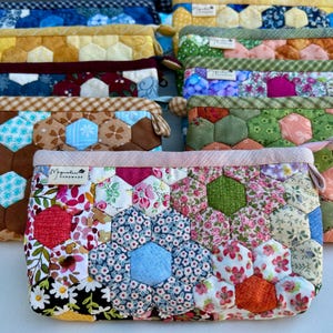 May include: A collection of handmade fabric pouches with a patchwork design. The pouches feature a variety of floral and geometric patterns in shades of blue, green, yellow, and brown. Each pouch has a zipper closure and a small tag that reads "Magnolia Handmade".