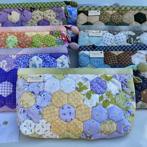 May include: A collection of rectangular zippered pouches featuring a quilted hexagon patchwork design in various floral and geometric patterns. Each pouch has a small fabric tag labeled Magnolia Handmade and a green fabric trim along the top edge.